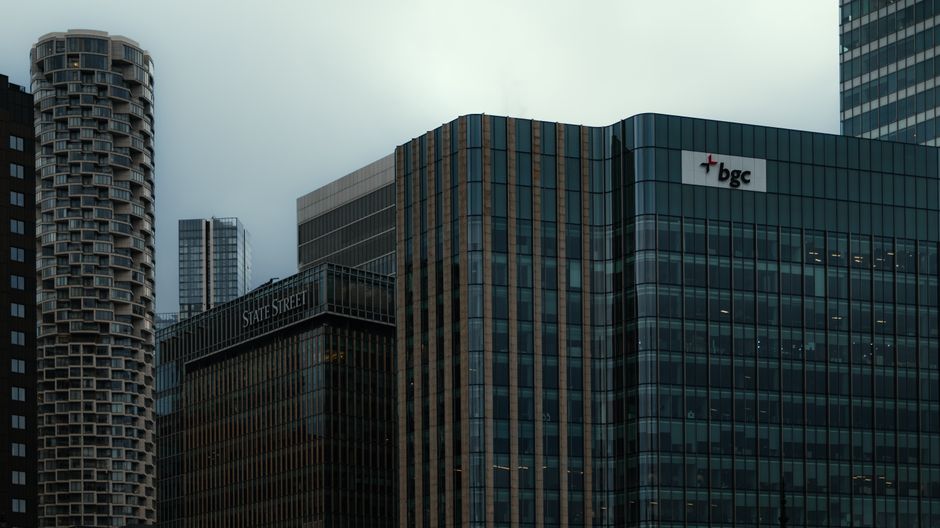 A panoramic view of the Canary Wharf financial district in London during dusk, featuring tall skyscrapers with illuminated windows, including the distinctive twisting building on the right, along with