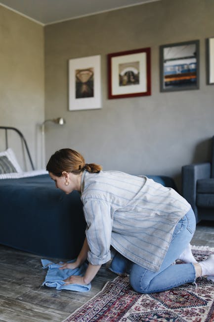 A woman with brown hair tied back, dressed in a light blue striped shirt and jeans, is kneeling on a patterned area rug while cleaning a dark blue upholstered bed frame with a blue cloth in a bedroom.